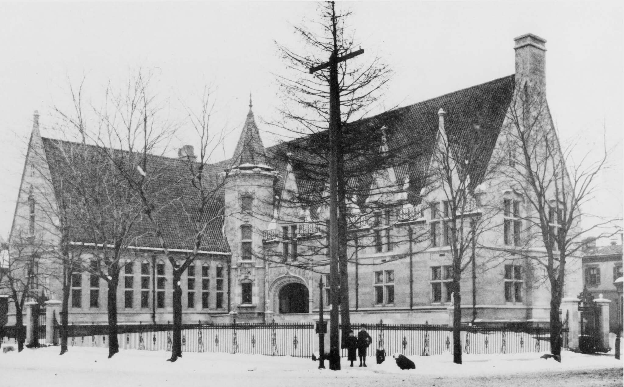 A winter photograph of the library around 1900: Gothic limestone gables, a corner tower and steep slate roof above an iron fence, with bare young trees on the snowy lawn.