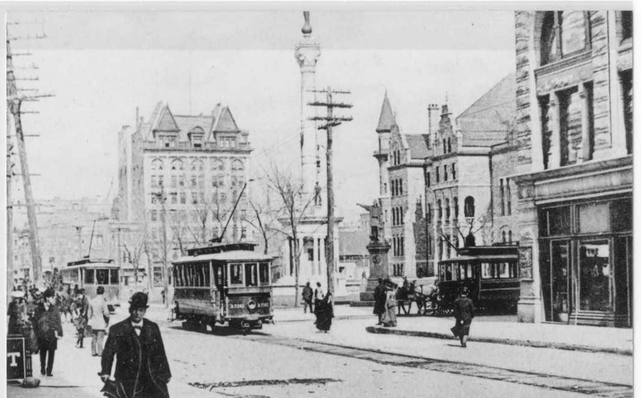 A photograph of the Square around 1905: open trolley cars crossing in front of the memorial column, pedestrians in hats and long coats, the courthouse behind bare trees.