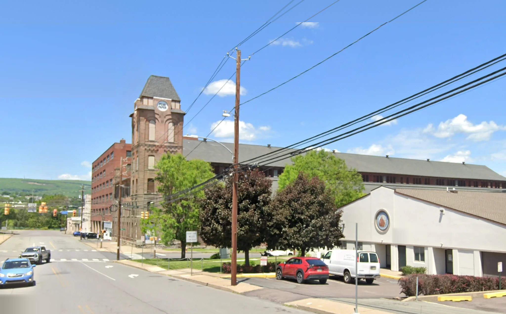 The site today: the same brick tower, its spire gone and a PENN PAPER sign across its face, standing over a long low shop building, with a modern one-storey warehouse and a parking lot filling the corner in front.