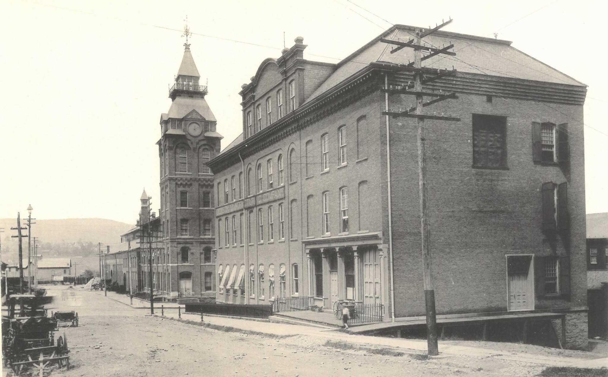 The Dickson works in the 1890s: a tall brick clock tower with a steep spire, roof balcony and weathervane rising over the shops, and a three-storey brick office block on the corner with awnings, a columned entrance porch and the company name lettered across its front, above a dirt street with wagons standing at the left.