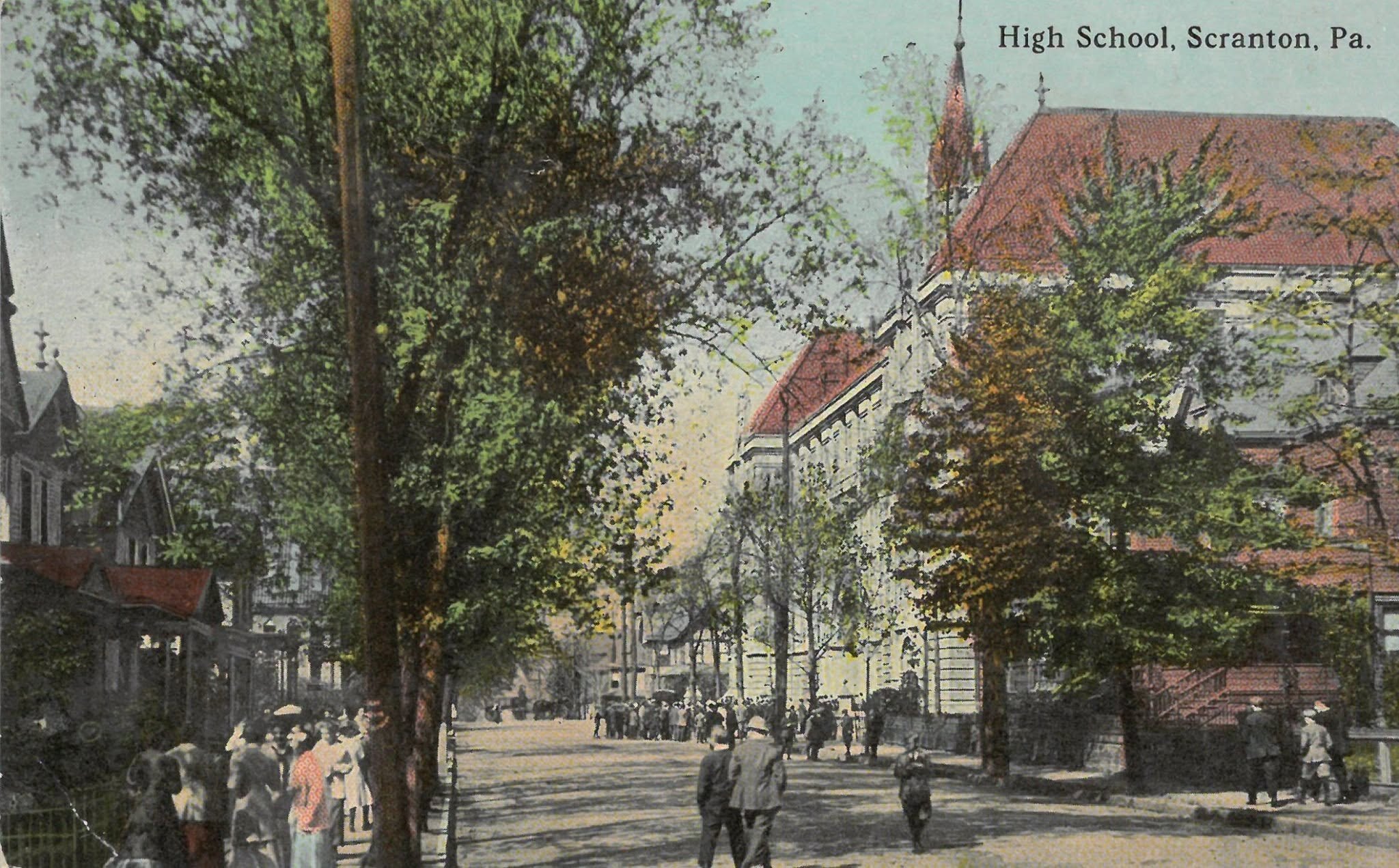 A hand-coloured 1910 postcard of the same block: elms arch over a crowded sidewalk as a crowd streams toward the school entrance.
