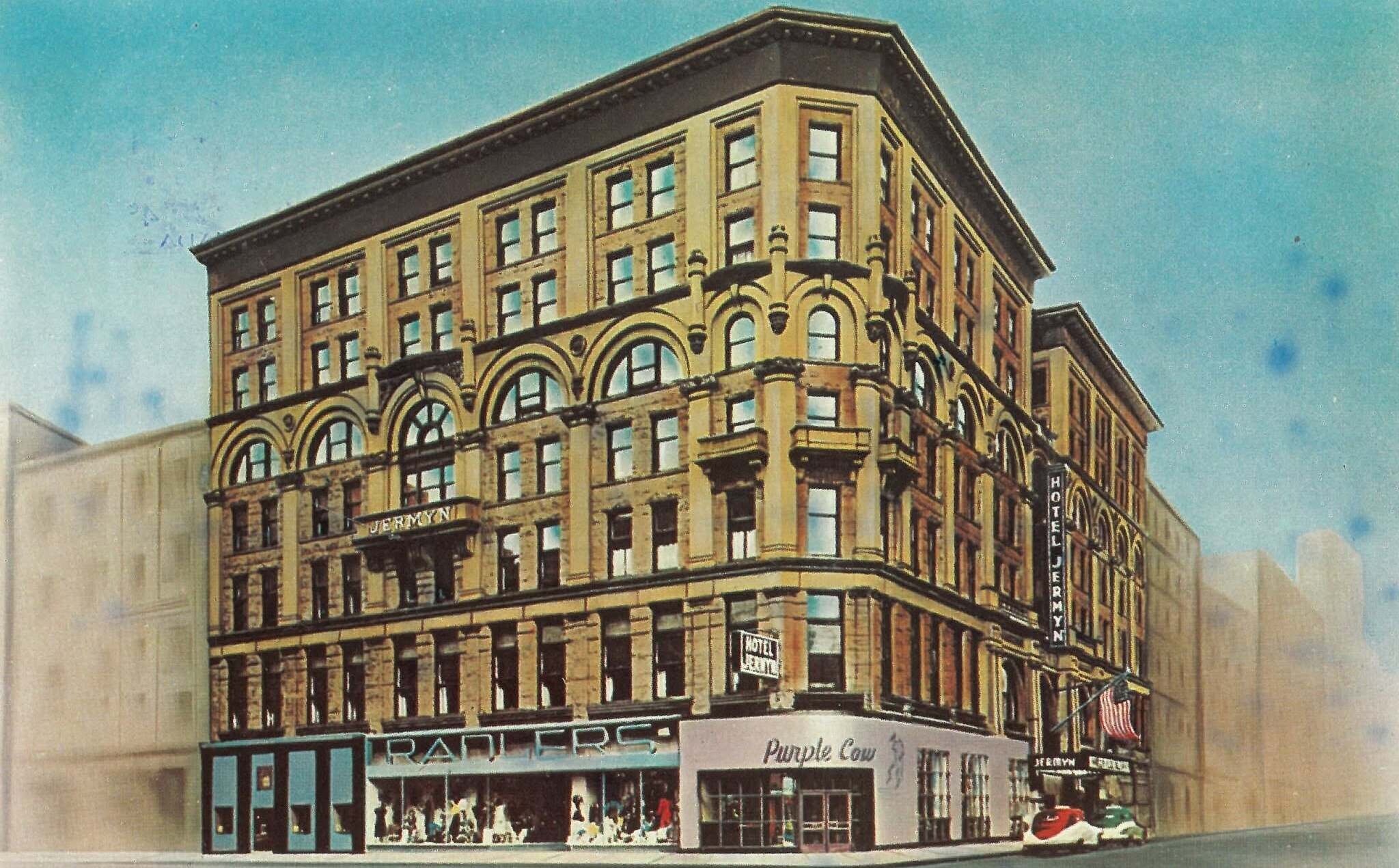 A tinted 1950s postcard of the Hotel Jermyn: a six-storey corner block with arched windows, a vertical HOTEL JERMYN sign, and Radler's and the Purple Cow lettered across the shopfronts below.