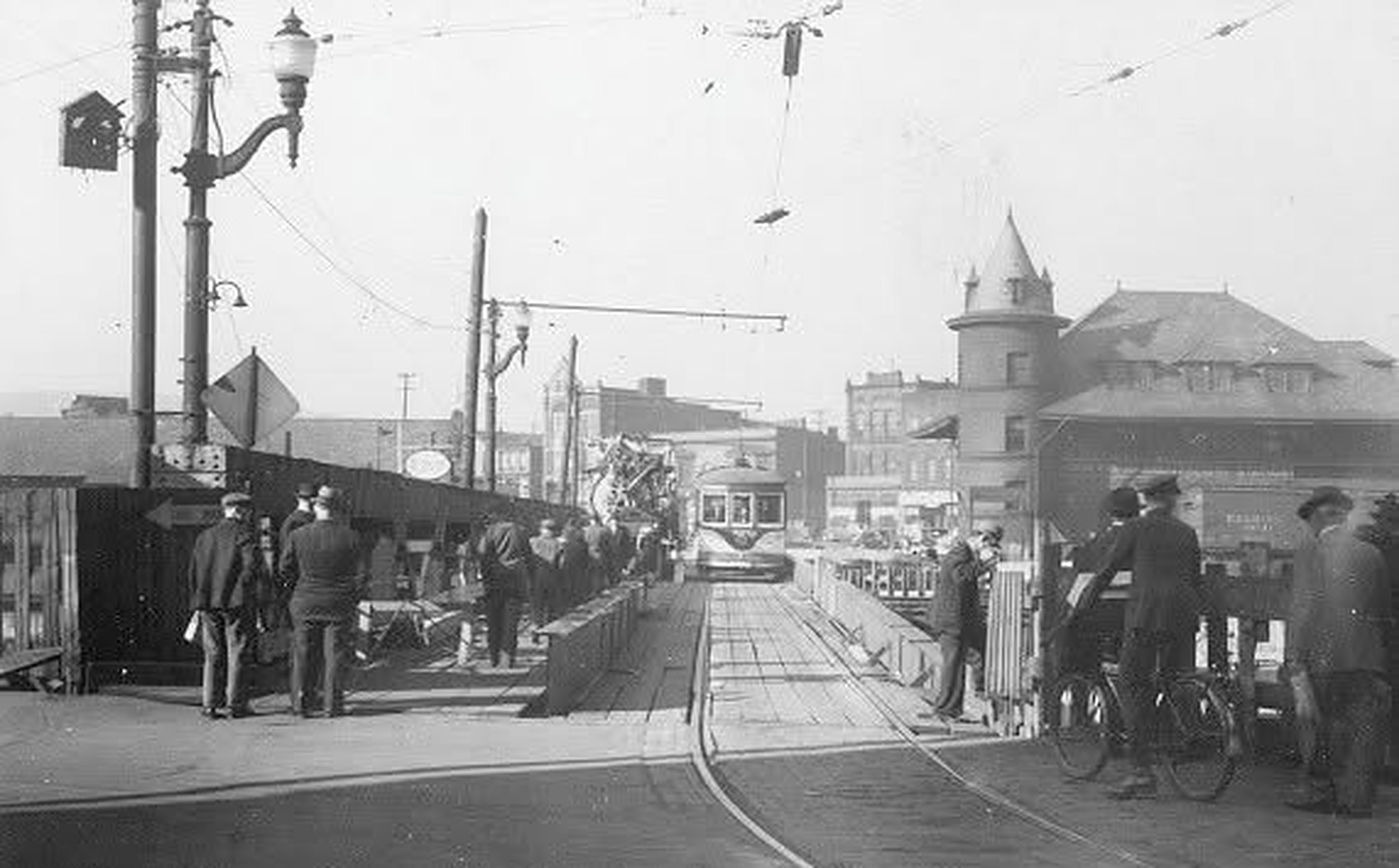 The bridge in the early 1900s: men in flat caps and overcoats standing along a plank barrier on the left, trolley tracks running up the deck toward a streetcar in the middle distance, overhead wires strung from poles, an ornate cast-iron lamp standard and a diamond warning sign at the left edge, and men with bicycles at the right-hand rail. The station's turret rises above the roofs on the right.