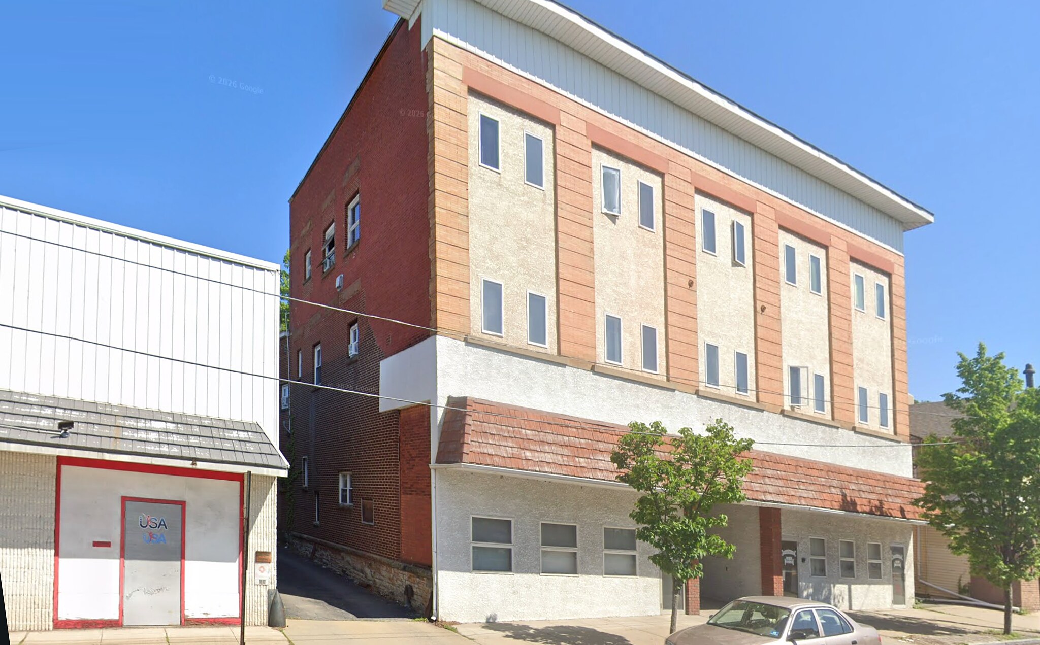 The same corner today: the building re-clad in pale stucco and metal panel, its cornice removed, a car parked at the kerb.