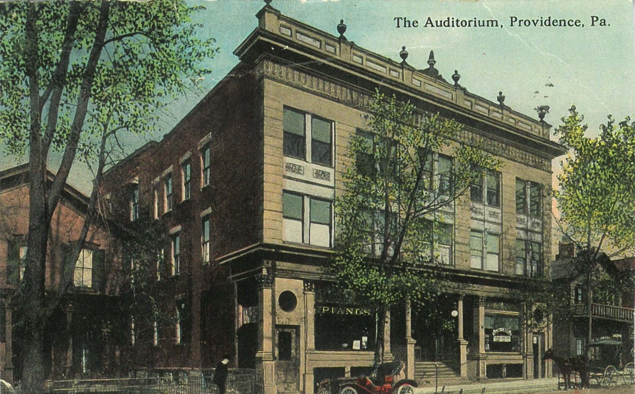 A postcard of the Auditorium around 1910: a three-storey corner block with a heavy cornice topped by stone urns, a piano dealer at street level, and a horse and cart at the kerb.