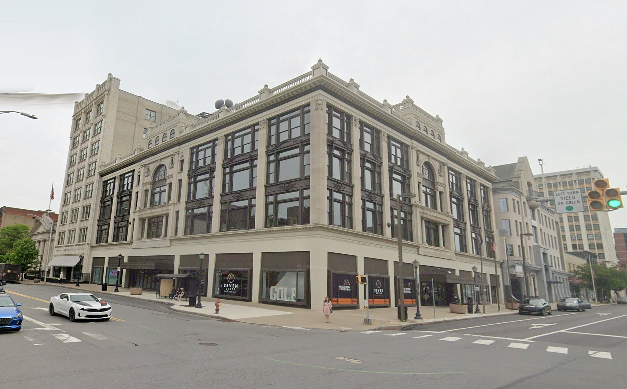 The same corner today: the department store building intact under a pale overcast sky, its roof signs gone, cars waiting at the crossing.