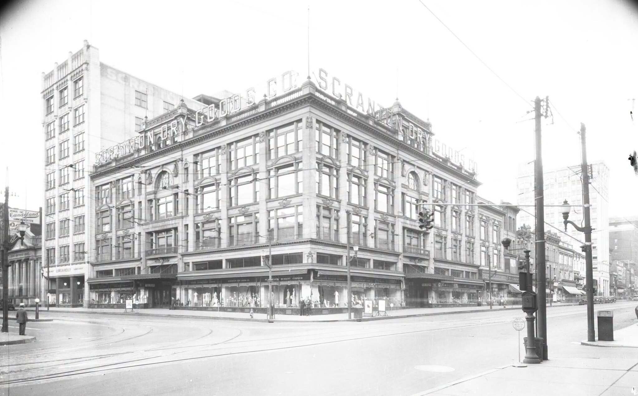A 1920s glass plate negative of the corner: a five-storey department store with SCRANTON DRY GOODS CO. lettered across the roofline, display windows below, streetcar tracks curving through the empty intersection.