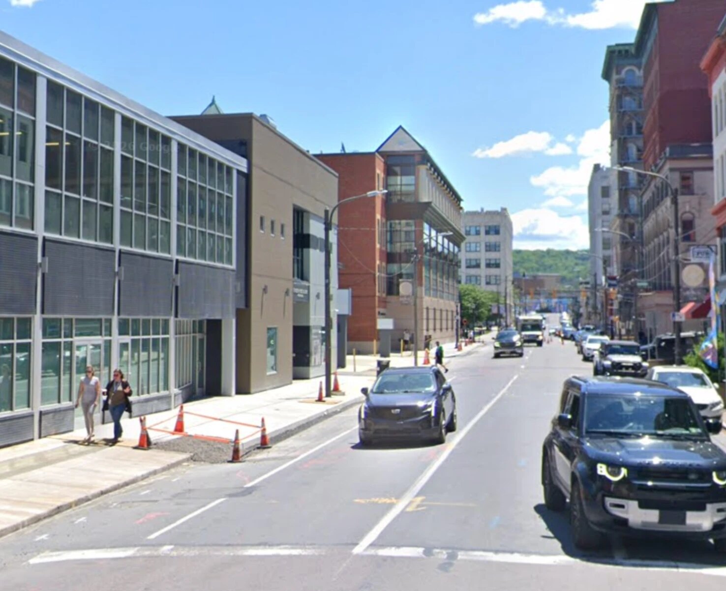 Spruce Street today: a glass-fronted modern building on the left where the old storefronts stood, cars in the travel lanes where the streetcar tracks ran, and the same ornate brick building still standing on the right in the distance.