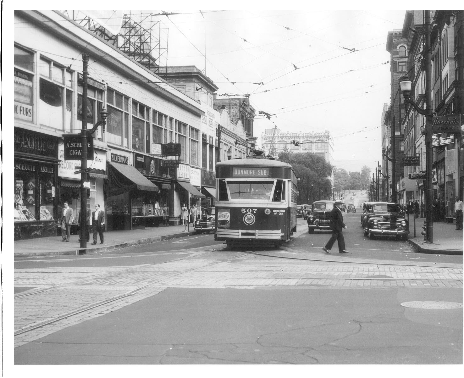 A streetcar reading DUNMORE SUB crosses the 400 block of Spruce Street in 1950, overhead wires crisscrossing above it, with a man crossing in front of it and storefronts, awnings and parked cars lining both sides of the street.