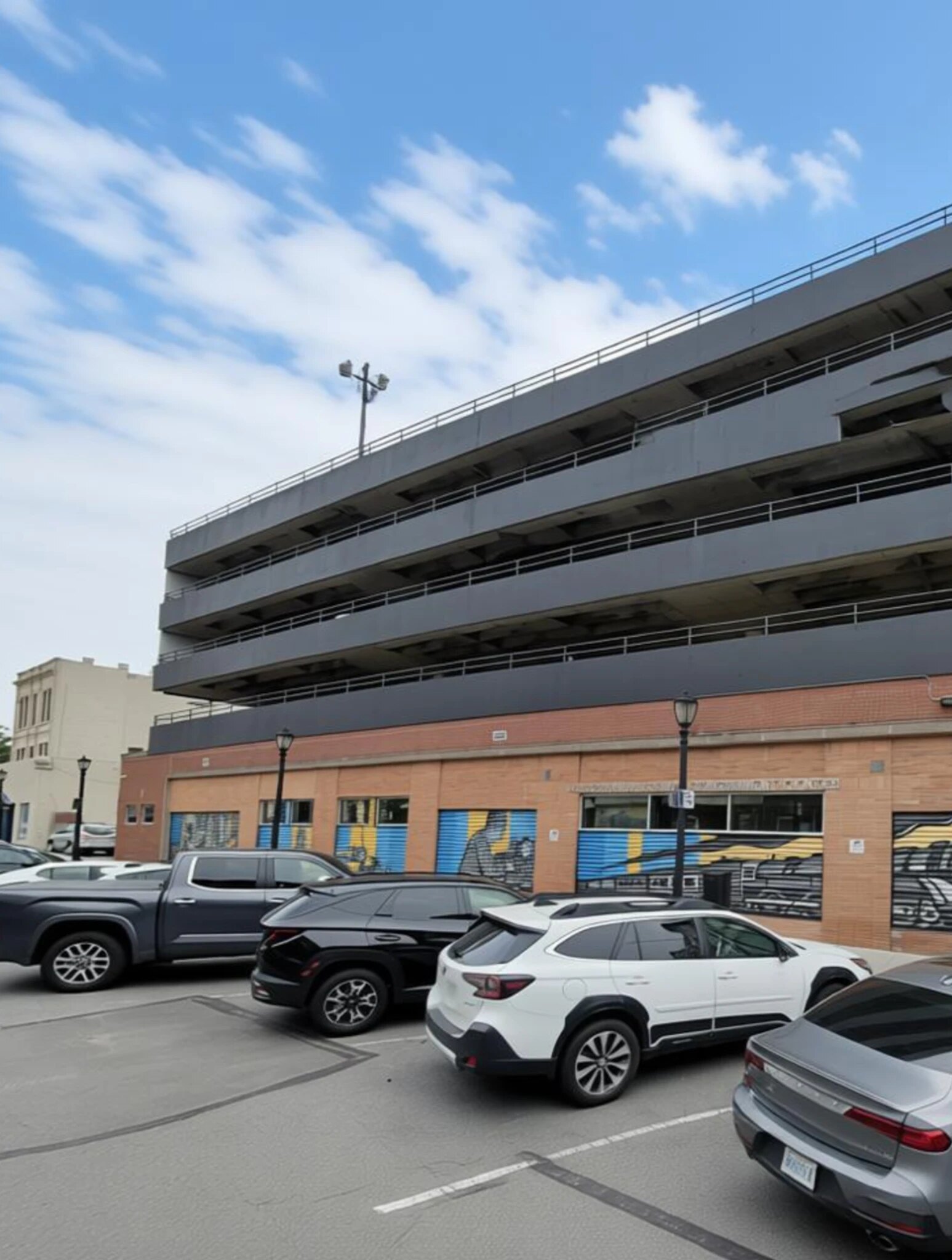 The site today: a multi-storey concrete parking garage rises over a low brick storefront wrapped in a mural, with a parking lot filling the corner in front.