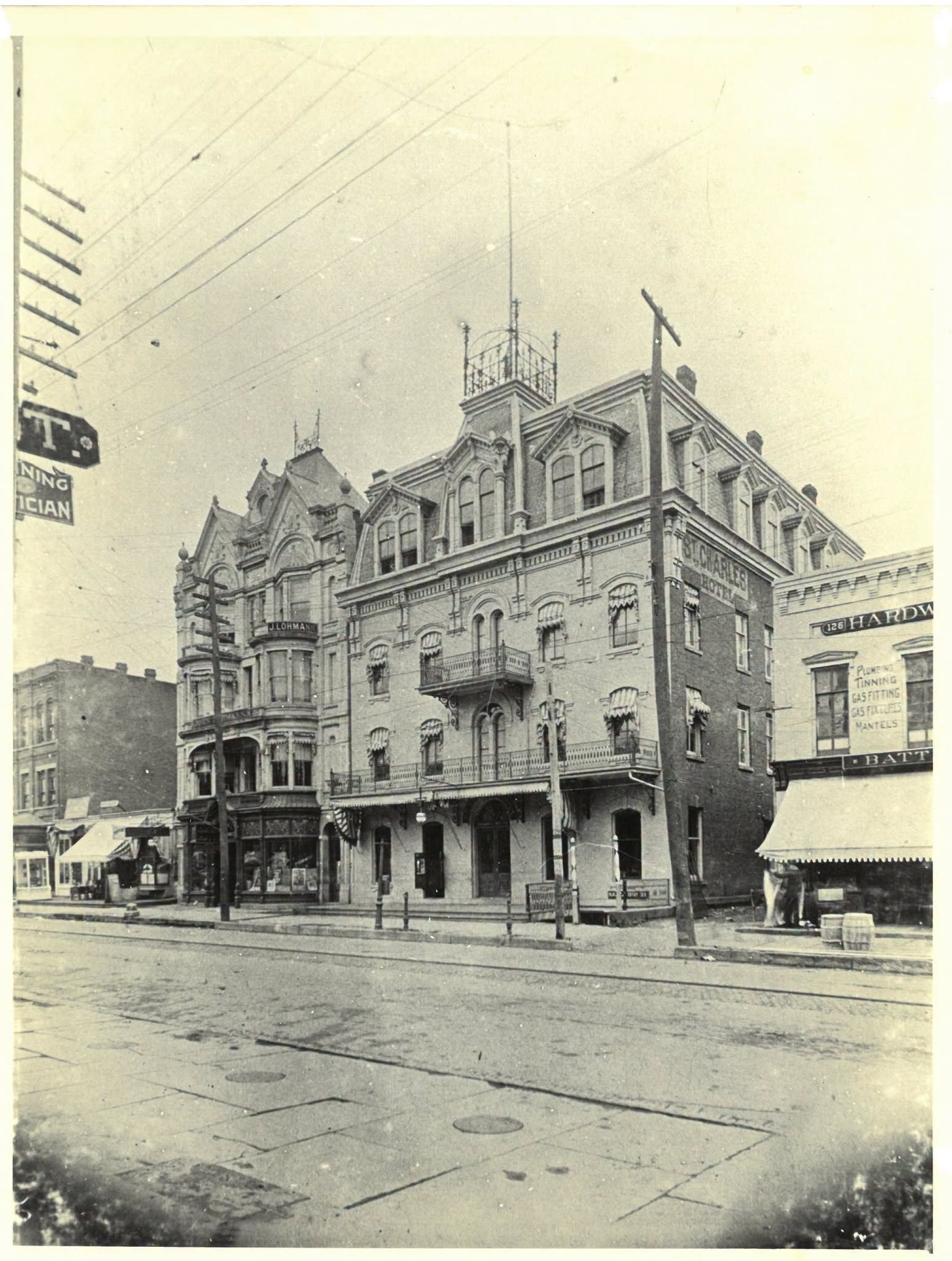 The St. Charles Hotel around 1900: a four-storey brick hotel with a mansard roof, iron balconies and awnings at every floor, a rooftop widow's walk, and the ornate gabled front of the Conway House next door, seen across a street with trolley tracks.