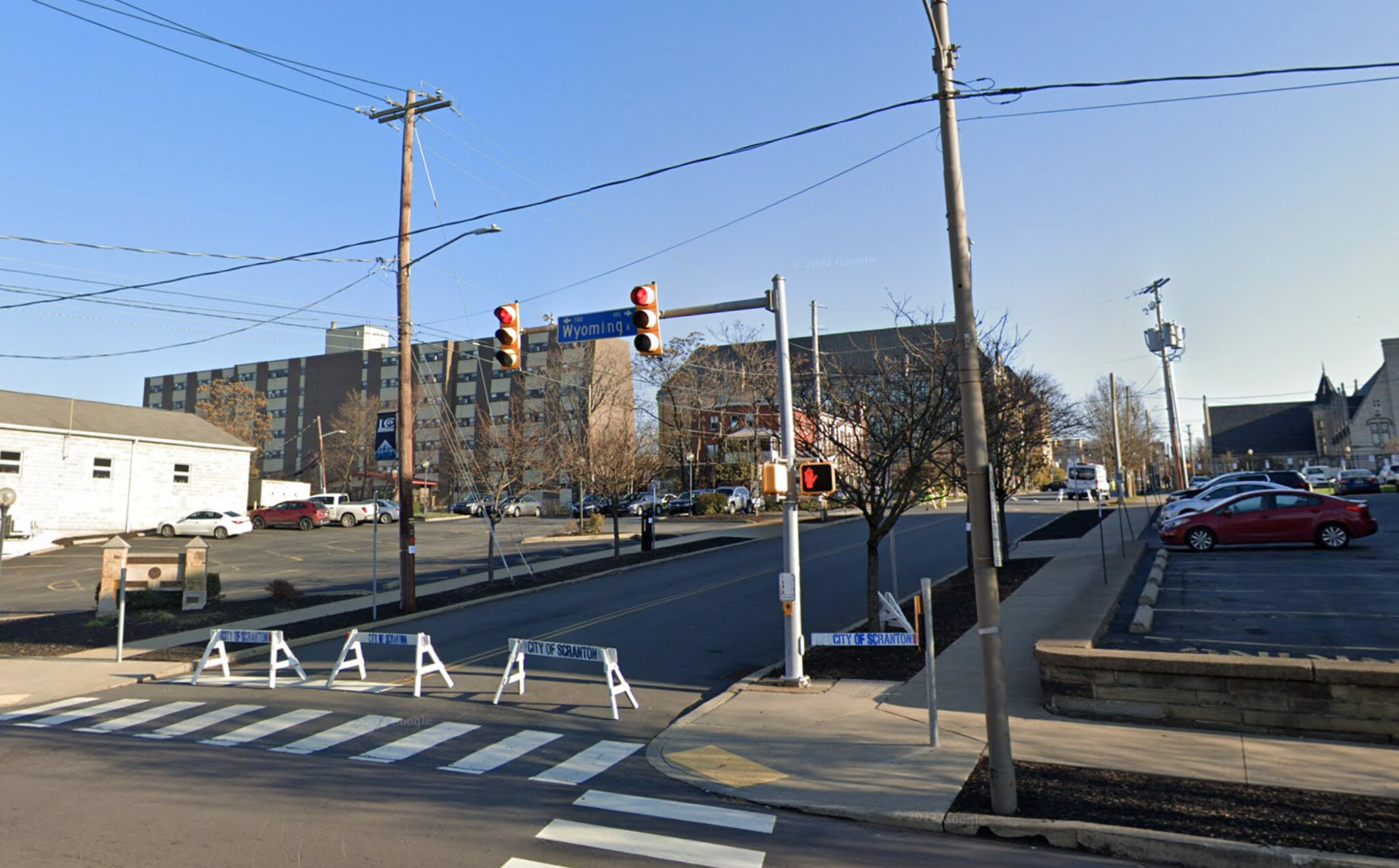 The corner today: the hotel site is a flat parking lot behind barricades, with a traffic signal at the crossing and the stone retaining wall still holding the raised ground on the right.