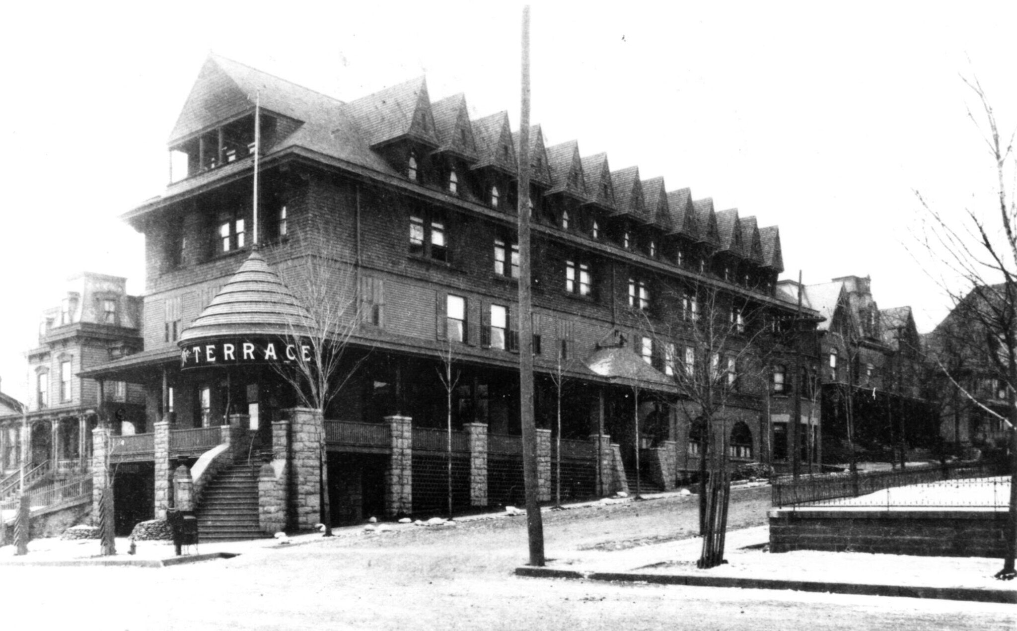 A winter photograph of the Hotel Terrace: a four-storey shingled hotel with dormers along its roof and a conical corner turret lettered TERRACE, standing above a stone terrace wall on the corner.