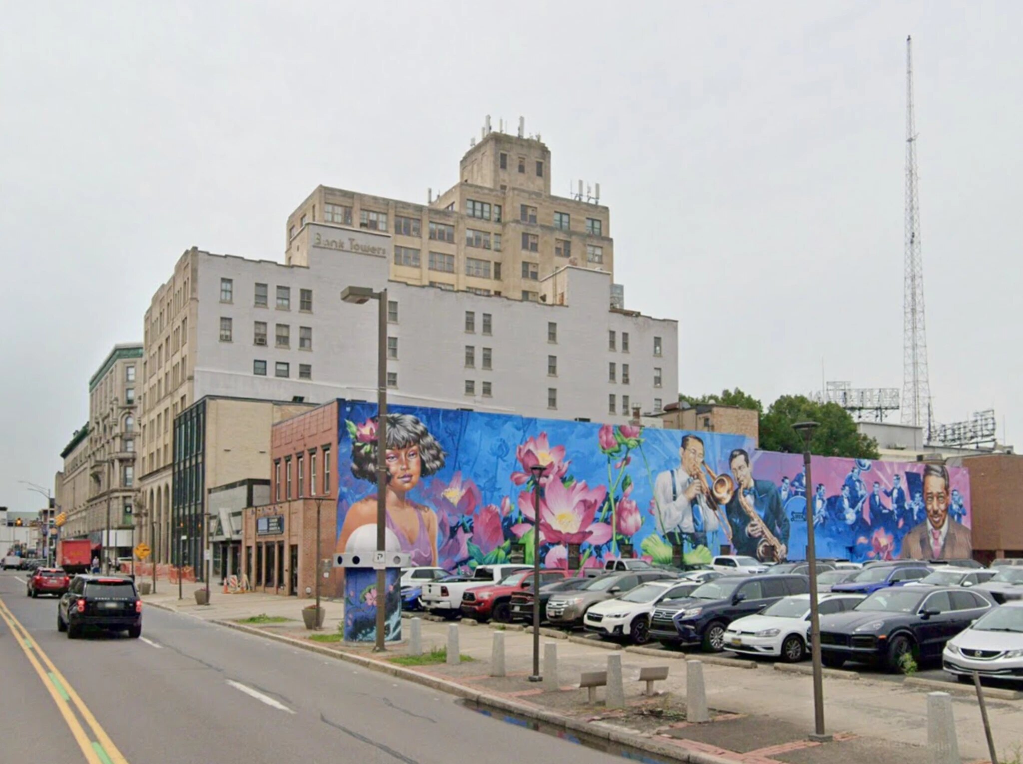 The block today: a plain white and tan tower called Bank Towers rises behind a low brick building and a long wall painted with a mural of flowers and jazz musicians, over a surface parking lot.
