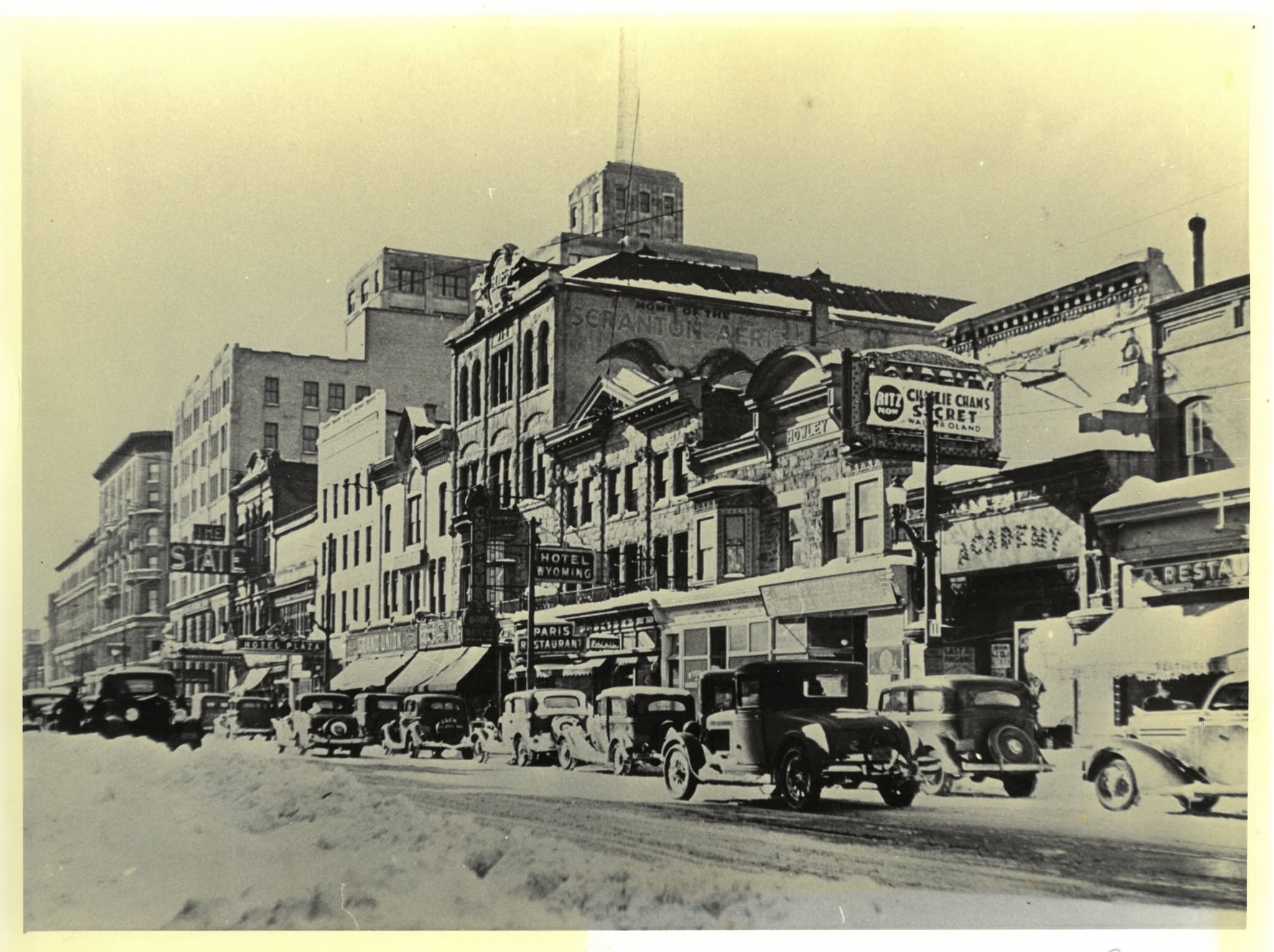 Wyoming Avenue under snow in 1936: a row of theater and restaurant buildings lit with signs for the State, the Academy and the Ritz, whose marquee advertises Charlie Chan's Secret, with a stepped tower rising behind them and cars parked nose-in along the curb.